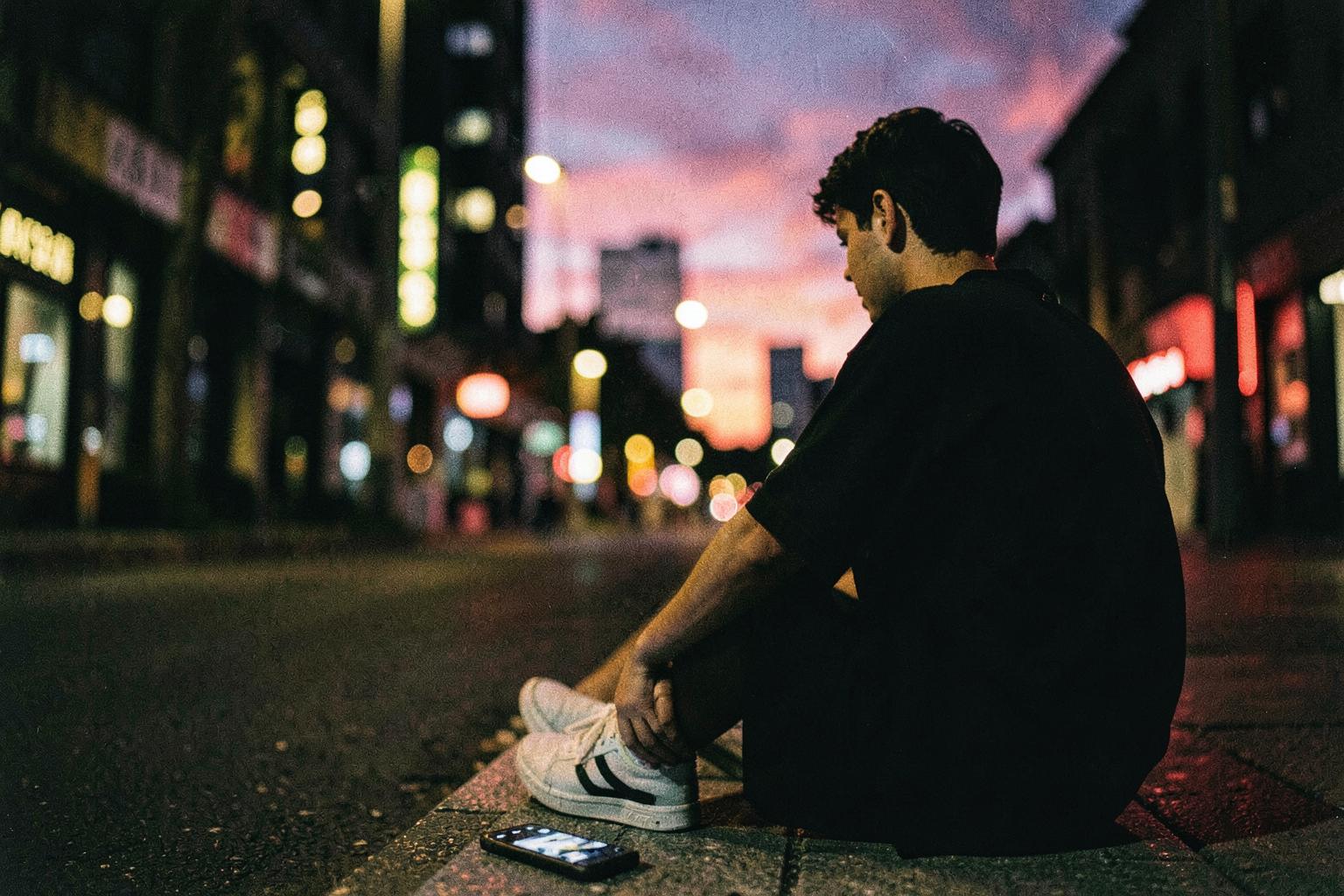 A person sitting on a sidewalk at night, phone on the ground, city neon glowing in front of them
